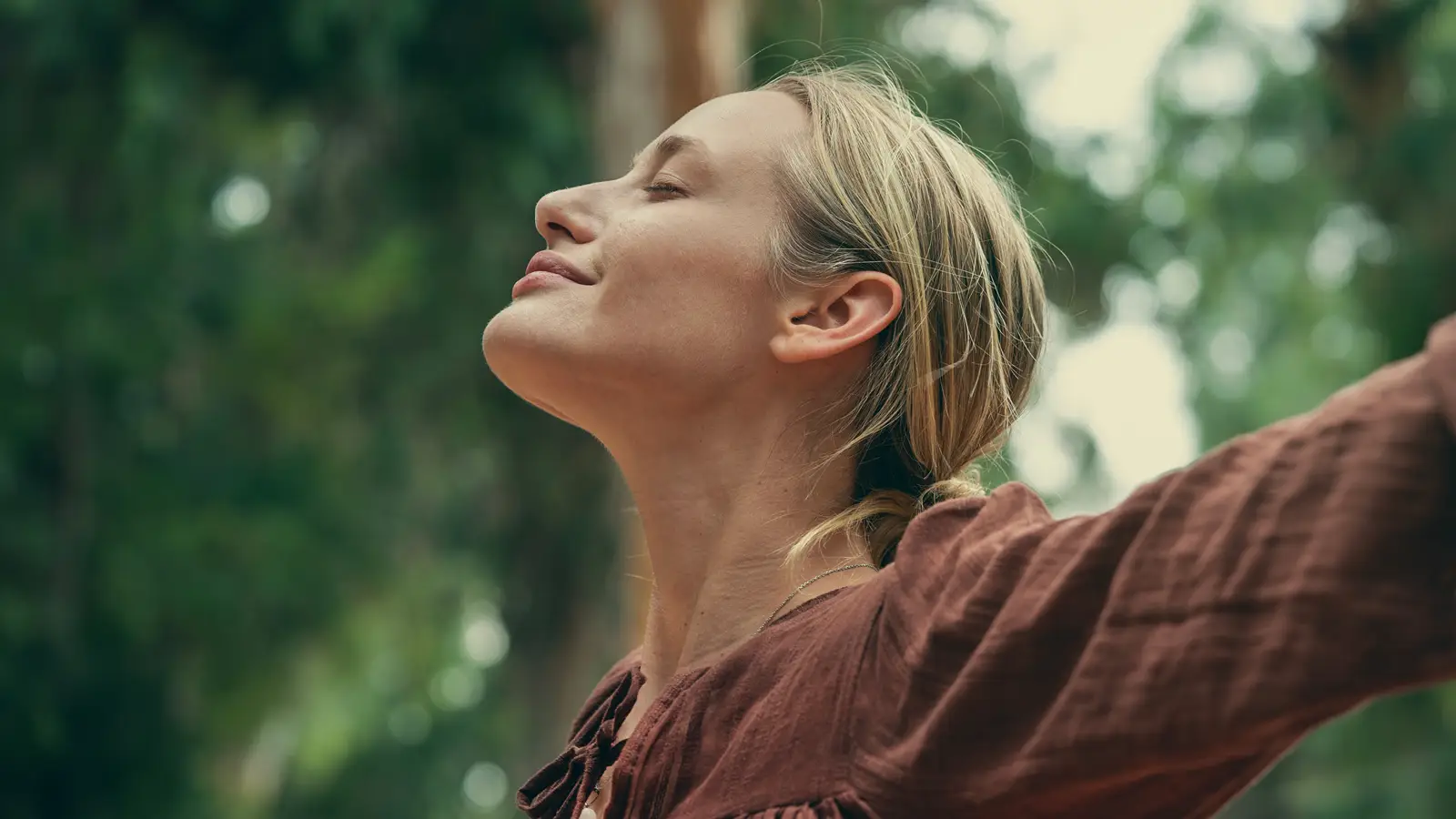 A lady, with her eyes closed and face, facing the sky, smiling with her arms spread wide in a forest.